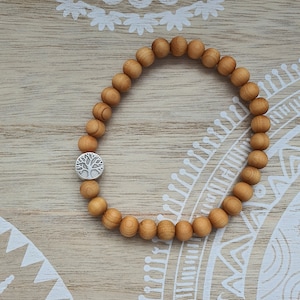 Könnte beinhalten: Ein Holzperlenarmband mit einem silbernen Lebensbaum-Anhänger. Das Armband ist braun und die Perlen sind rund. Der Anhänger befindet sich in der Mitte des Armbands.