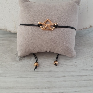 Könnte beinhalten: Ein verstellbares Armband aus schwarzem Kordel mit einem goldenen Papierboot-Anhänger. Das Armband wird auf einem grauen Samtkissen präsentiert.