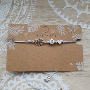 Könnte beinhalten: Ein weißes elastisches Armband mit silbernen Anhängern, darunter ein Pfotenabdruck, ein Buchstabe "S" und ein Herz. Das Armband wird auf einer braunen Karte mit dem Text "Musthave" präsentiert.