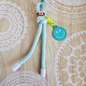 Könnte beinhalten: Ein hellblaues Schlüsselband mit einem Smiley-Anhänger. Das Schlüsselband hat ein geknotetes Design mit weißen Spitzen und einem grünen Verschluss. Ein kleines Etikett mit einem Smiley ist angebracht. Der Hintergrund ist eine helle Holzoberfläche mit weißen kreisförmigen Mustern.