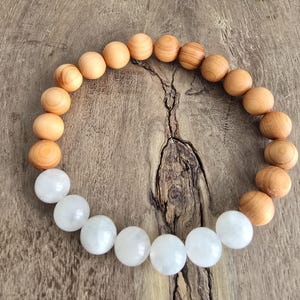 Könnte beinhalten: Ein Armband mit abwechselnd hellbraunen Holzperlen und weißen Edelsteinperlen. Die Perlen werden auf einer Holzoberfläche präsentiert. Das Design des Armbands hebt den Kontrast zwischen den natürlichen Materialien hervor.