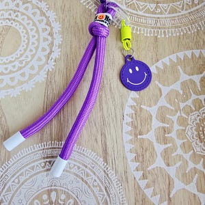 Könnte beinhalten: Ein lila Schlüsselband mit einem Smiley-Anhänger. Das Schlüsselband hat ein geknotetes Design mit zwei lila Schnüren, die in weißen Spitzen enden. Ein kleiner, runder, lila Anhänger mit einem weißen Smiley-Gesicht ist befestigt. Es hat auch einen Metallverschluss und eine gelbe Spirale.