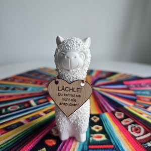 Könnte beinhalten: Eine weiße Keramik-Alpaka-Figur mit strukturiertem Fell, die ein herzförmiges Holzschild hält. Auf dem Schild steht "LÄCHLE! Du kannst sie nicht alle anspucken!". Das Alpaka steht auf einem bunten, gemusterten Textil.