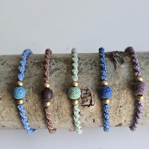 Könnte beinhalten: Fünf handgefertigte Armbänder mit farbigen Lavasteinperlen. Jedes Armband hat eine andere Farbe des geflochtenen Kordels: blau, braun, grün, dunkelblau und lila. Die Perlen sind mit goldfarbenen Metallperlen akzentuiert.