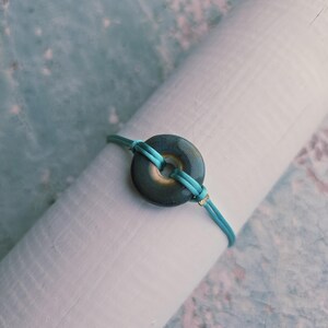 Könnte beinhalten: Ein türkisfarbenes Armband mit einem dunkelgrünen, kreisförmigen Mittelstück. Das Armband besteht aus einer türkisfarbenen Kordel und hat einen kleinen goldenen Akzent. Das Armband wird auf einem weißen zylindrischen Objekt vor einem hellblau-weißen Hintergrund präsentiert.