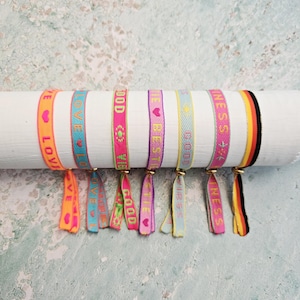 Könnte beinhalten: Eine Sammlung farbenfroher gewebter Armbänder mit positiven Wörtern wie "LOVE", "GOOD" und "VIBES". Jedes Armband hat ein anderes Farbschema mit Text und kleinen Herz- oder Blumengrafiken. Die Armbänder werden auf einem weißen zylindrischen Objekt präsentiert.