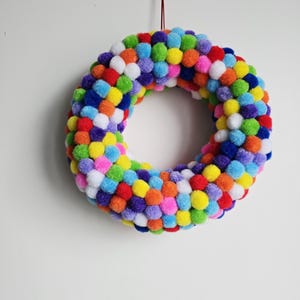 Könnte beinhalten: Ein farbenfroher Kranz aus kleinen, flauschigen Pompons in verschiedenen Farben, darunter Rot, Orange, Gelb, Grün, Blau, Lila und Weiß. Der runde Kranz hängt an einem dünnen roten Faden vor einem weißen Hintergrund.