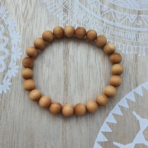 Könnte beinhalten: Ein Armband aus hellbraunen Holzperlen. Die runden Perlen sind poliert. Das Armband ist kreisförmig angeordnet. Es liegt auf einer hellbraunen Holzoberfläche mit einem weißen Dekorationsmuster.