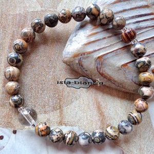 Könnte beinhalten: Ein Perlenarmband mit runden, gemusterten Steinen in Braun-, Beige-, Grau- und Schwarztönen, unterbrochen von einer einzelnen transparenten Perle. Der Text "isla-blanca" ist in der Mitte der Armbandschleife deutlich zu erkennen.