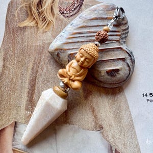 Könnte beinhalten: Ein dekorativer Anhänger mit einer geschnitzten hellbraunen Buddha-Figur in meditativer Sitzhaltung, gekrönt von einer kleinen texturierten braunen Perle und einem silberfarbenen Verschluss, endend in einer glatten, cremeweißen Kristallspitze.
