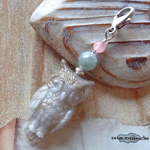 Könnte beinhalten: Ein detaillierter Eulen-Anhänger mit einem silbernen Verschluss, rosa und grünen Perlen. Die Eule hat eine helle, perlmuttartige Farbe mit aufwendigen Schnitzereien. Der Anhänger ist an einem silbernen Verschluss befestigt.