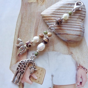 Könnte beinhalten: Ein silbernes Charm-Armband mit einer Giraffe und einem kleinen Tieranhänger. Das Armband hat eine Mischung aus Perlen in Braun-, Grün- und Weißtönen, darunter Perlen. Der Anhänger ist an einem silbernen Verschluss befestigt.