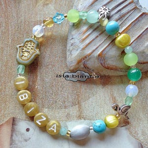 Könnte beinhalten: Ein Armband mit bunten Perlen, auf dem das Wort "Ibiza" in goldenen Perlen geschrieben steht. Das Armband enthält türkisfarbene, gelbe, grüne und transparente Perlen. Ein Hamsa-Hand-Anhänger und ein Flamingo-Anhänger sind ebenfalls enthalten.