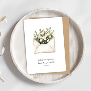 Könnte beinhalten: Eine weiße Grußkarte mit einer Aquarellillustration von Blumen, die aus einem Umschlag quellen. Die Karte liegt auf einem Holztablett, dahinter ein brauner Umschlag. Der Text auf der Karte lautet "Alle Dinge sind möglich dem, der da glaubt. Markus 9,23."