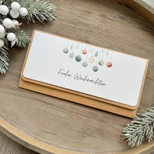 Könnte beinhalten: Eine weiße Karte mit einem braunen Umschlag. Die Karte zeigt eine Aquarell-Illustration von Weihnachtskugeln, die an einer Schnur hängen. Der Text "Frohe Weihnachten" ist in schwarzer Kursivschrift auf der Karte geschrieben.