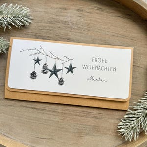 Könnte beinhalten: Eine weiße Karte mit einem braunen Umschlag. Die Karte zeigt eine Zeichnung eines Zweigs mit schwarzen Sternen und Tannenzapfen, die daran hängen. Der Text auf der Karte lautet "Frohe Weihnachten" und "Martin".