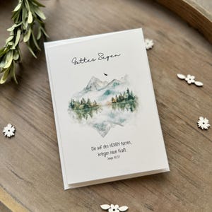 Könnte beinhalten: Eine weiße Karte mit einer Aquarell-Illustration von Bergen und einem See. Der Text "Gottes Segen" befindet sich oben auf der Karte. Der Text "Die auf den HERRN harren, kriegen neue Kraft" befindet sich unten auf der Karte. Der Text "Jesaja 40,31" befindet sich unter dem Zitat.
