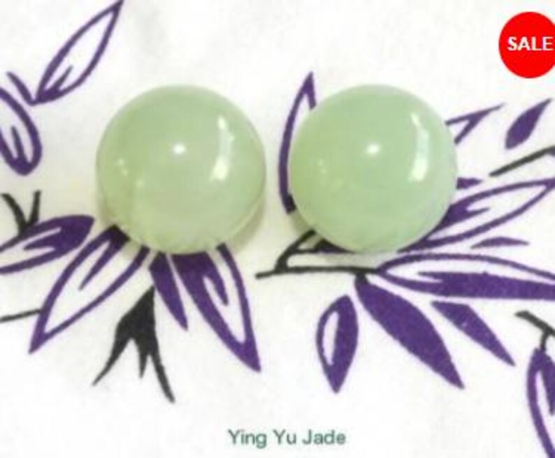 May include: Two pale green, round jade spheres. The spheres are on a white cloth with a purple leaf pattern. A red circle with the word "SALE" is in the upper right corner. The text "Ying Yu Jade" is at the bottom.