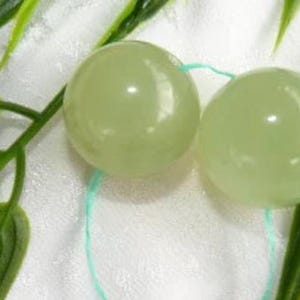 May include: Two pale green, spherical beads connected by a light blue string. The beads are smooth and glossy, reflecting light. Green leaves and stems are in the background, creating a natural aesthetic.