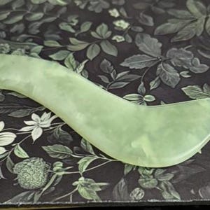 May include: A light green jade gua sha tool with a curved, ergonomic design. The tool is smooth and polished, and it rests on a dark background with a floral pattern. This skincare tool is used for facial massage.