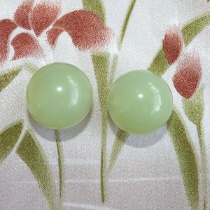 May include: Two pale green, round, glossy beads. The beads are set against a patterned background of white fabric with red and green floral designs. The beads are smooth and spherical.