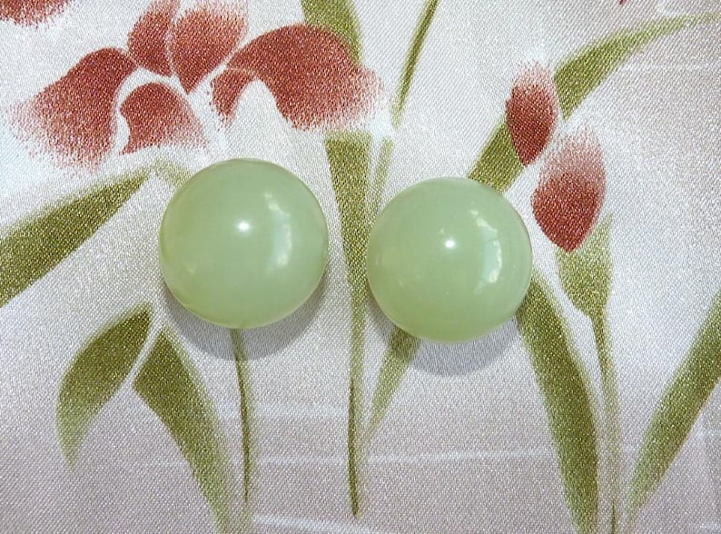 May include: Two pale green, round, glossy beads. The beads are set against a patterned background of white fabric with red and green floral designs. The beads are smooth and spherical.