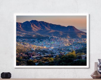 May include: A framed photograph of a city nestled in a valley, with mountains in the background. The city is a mix of buildings, trees, and roads. The sky is a soft pink and orange, with clouds in the distance.