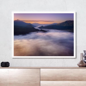 May include: A framed photograph of a mountain range with a layer of fog in the valley below. The sky is a gradient of purple, pink, and orange.