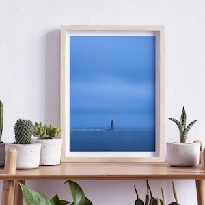 May include: A framed print featuring a lighthouse in the ocean under a blue sky. The artwork is in a light wood frame and is displayed on a wooden shelf with potted plants. The scene evokes a sense of calm and tranquility.