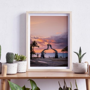 May include: A framed photograph of a sunset over the ocean with a large sculpture of two hands reaching towards each other in the foreground.