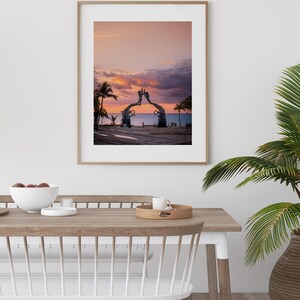 May include: A framed photograph of a sunset over the ocean with a large metal sculpture in the foreground. The sculpture is shaped like two hands reaching up towards the sky. The sky is a vibrant pink and orange color, and the water is a deep blue.