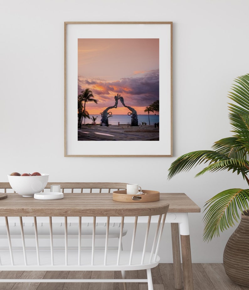May include: A framed photograph of a sunset over the ocean with a large metal sculpture in the foreground. The sculpture is shaped like two hands reaching up towards the sky. The sky is a vibrant pink and orange color, and the water is a deep blue.