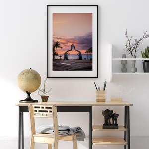 May include: A framed photograph of a sunset over the ocean with a large sculpture of two hands reaching towards each other in the foreground.