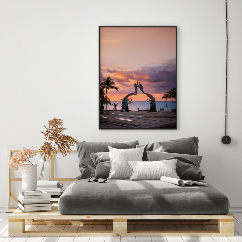 May include: A framed photograph of a sunset over the ocean with a large metal sculpture in the foreground. The sculpture is shaped like two hands reaching up towards the sky. The sky is a vibrant pink and orange, and the water is a deep blue.