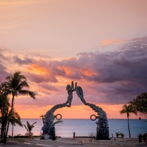 May include: A bronze sculpture of two figures reaching towards each other, forming an archway. The sculpture is set against a backdrop of a pink and orange sunset over a body of water.