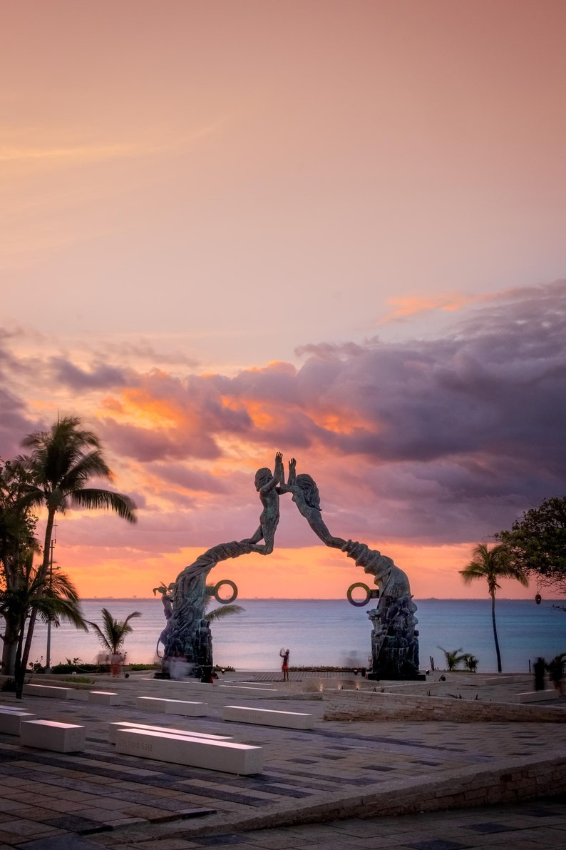May include: A bronze sculpture of two figures reaching towards each other, forming an archway. The sculpture is set against a backdrop of a pink and orange sunset over a body of water.