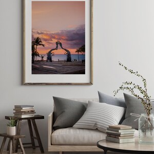 May include: A framed photograph of a metal sculpture of two hands reaching towards each other, set against a pink and purple sunset sky. The sculpture is located on a beach with palm trees and the ocean in the background.