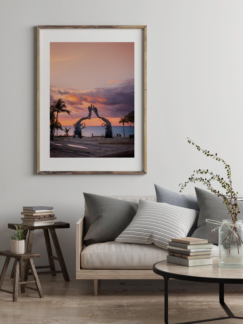 May include: A framed photograph of a metal sculpture of two hands reaching towards each other, set against a pink and purple sunset sky. The sculpture is located on a beach with palm trees and the ocean in the background.