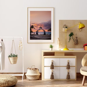 May include: A framed photograph of a sunset over the ocean with a large sculpture of two hands reaching towards each other in the foreground. The sculpture is made of metal and is painted in a light brown color. The photo is hanging on a white wall in a room with a wooden dresser and a plant.