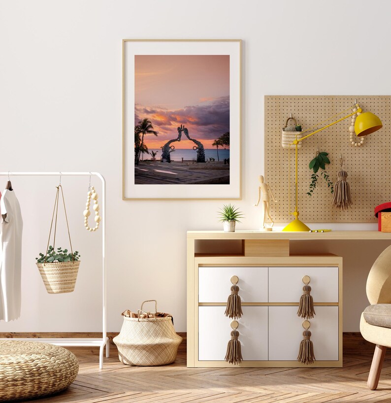 May include: A framed photograph of a sunset over the ocean with a large sculpture of two hands reaching towards each other in the foreground. The sculpture is made of metal and is painted in a light brown color. The photo is hanging on a white wall in a room with a wooden dresser and a plant.