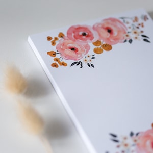 Könnte beinhalten: Ein weißer Notizblock mit floralem Design. Das Aquarelldesign zeigt rosa Rosen, kleine weiße Blüten und orangefarbene Akzente. Der Notizblock ist leer, mit dem floralen Design in der oberen linken und unteren rechten Ecke.
