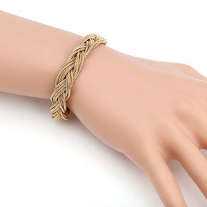 May include: A gold-colored braided bracelet. The bracelet is made of interwoven strands, creating a textured, woven appearance. The bracelet is shown on a person's wrist, showcasing its design and style. The bracelet is a fashion accessory.