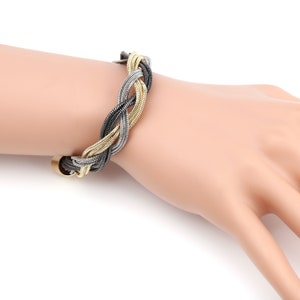 May include: Braided bracelet featuring interwoven strands of gold, silver, and black metal. The bracelet is displayed on a mannequin hand, showcasing its design and texture. The clasp is a gold-toned metal. This accessory is suitable for everyday wear or special occasions.