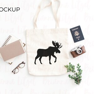 Puede incluir: Una bolsa de lona blanca con un dise&ntilde;o de silueta de alce negro. La bolsa est&aacute; colocada sobre una superficie blanca con un pasaporte, una c&aacute;mara y vegetaci&oacute;n.