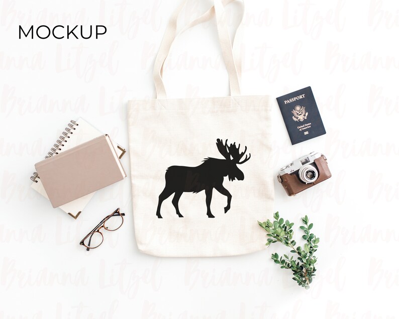 Puede incluir: Una bolsa de lona blanca con un dise&ntilde;o de silueta de alce negro. La bolsa est&aacute; colocada sobre una superficie blanca con un pasaporte, una c&aacute;mara y vegetaci&oacute;n.