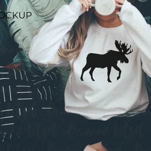 Puede incluir: Sudadera blanca con un gr&aacute;fico de silueta de alce negro. La sudadera la lleva puesta una persona y el fondo es un patr&oacute;n de hojas verdes.