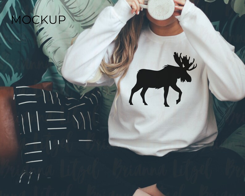 Puede incluir: Sudadera blanca con un gr&aacute;fico de silueta de alce negro. La sudadera la lleva puesta una persona y el fondo es un patr&oacute;n de hojas verdes.
