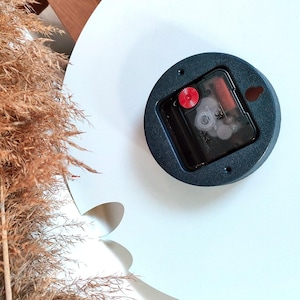 Peut inclure: Gros plan sur l'arri&egrave;re d'un m&eacute;canisme d'horloge. Le m&eacute;canisme est noir et poss&egrave;de un bouton rouge. L'horloge est mont&eacute;e sur une surface blanche.