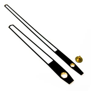 May include: Two black clock hands with white accents and gold metal attachments. The hands are on a white background.