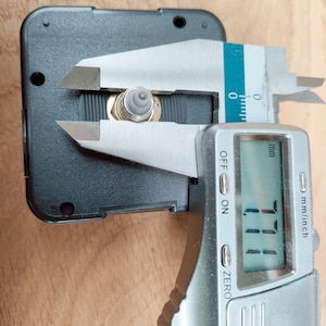 Puede incluir: Un calibre digital plateado mide un peque&ntilde;o mecanismo de reloj negro con una pieza de metal dorada. El calibre muestra 1,11 mm.